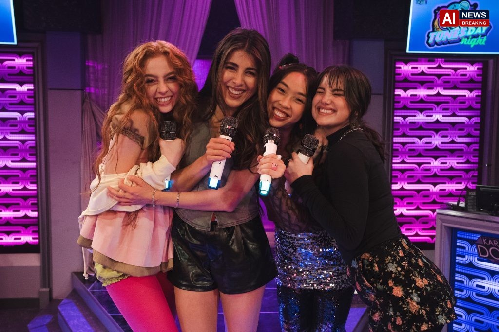 "Four young women smiling and hugging while holding microphones, standing together on a colorful karaoke stage decorated with purple lights and a "Tunes-day Night" sign, suggesting a lively and fun indoor environment."